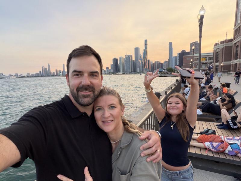 Greg and Natasha by the water at sunset with a city skyline behind them